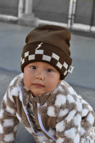 A baby wearing a brown and white checkered knit beanie with a small white lightning bolt logo, paired with a brown and white patterned fleece jacket, photographed outdoors.