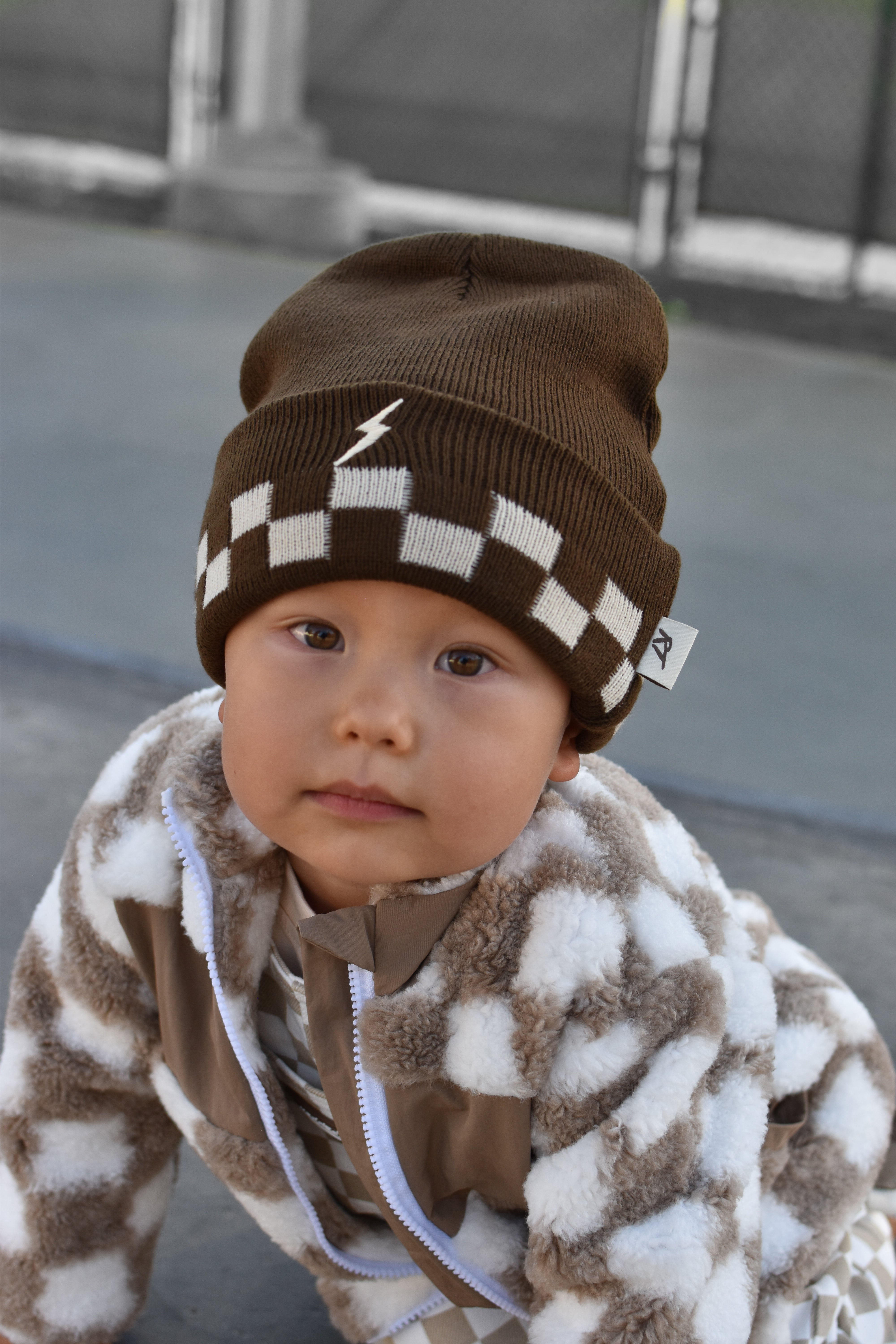 A baby wearing a brown and white checkered knit beanie with a small white lightning bolt logo, paired with a brown and white patterned fleece jacket, photographed outdoors.