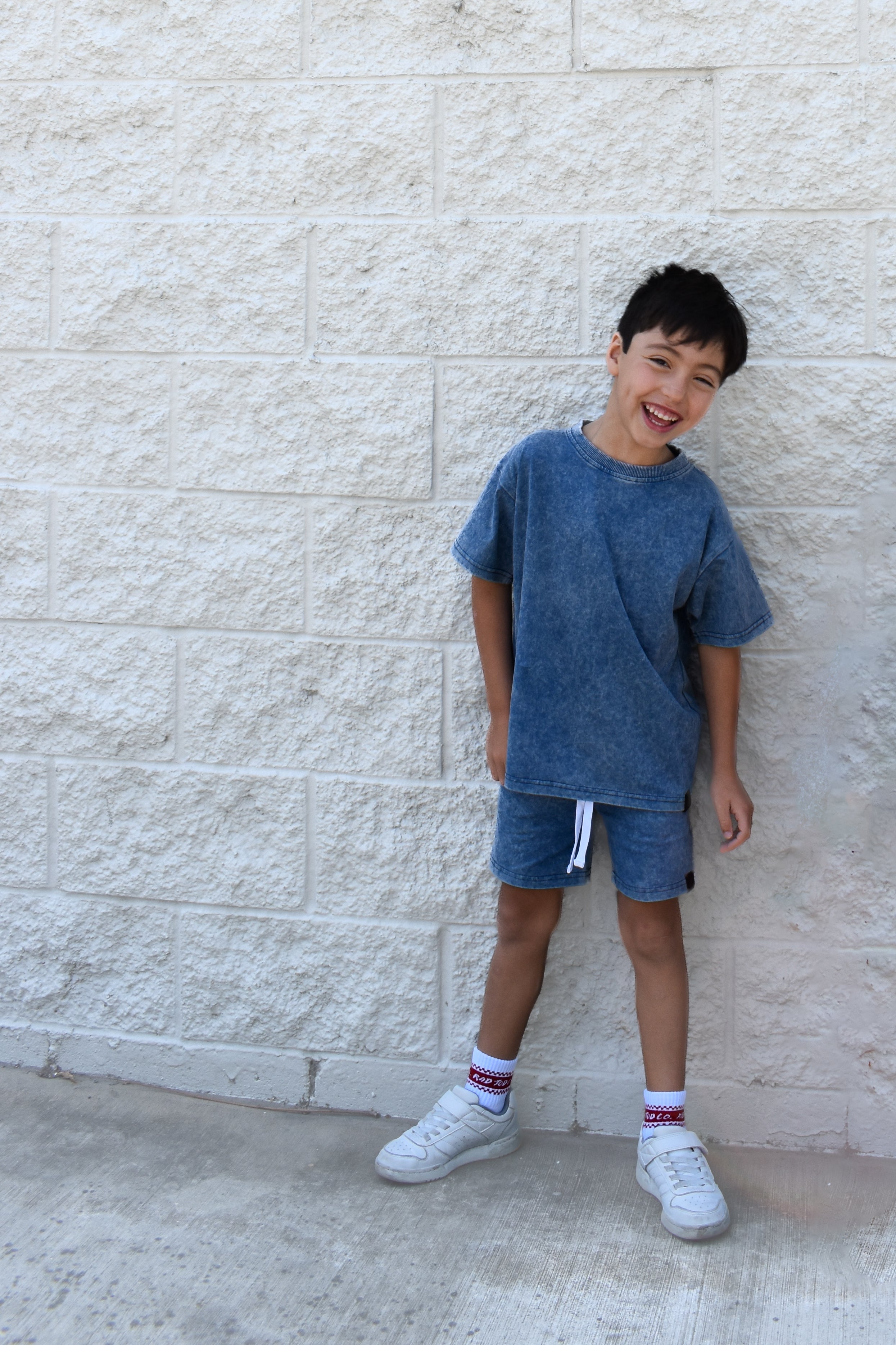 Child wearing a blue t-shirt and shorts standing against a light stone wall.