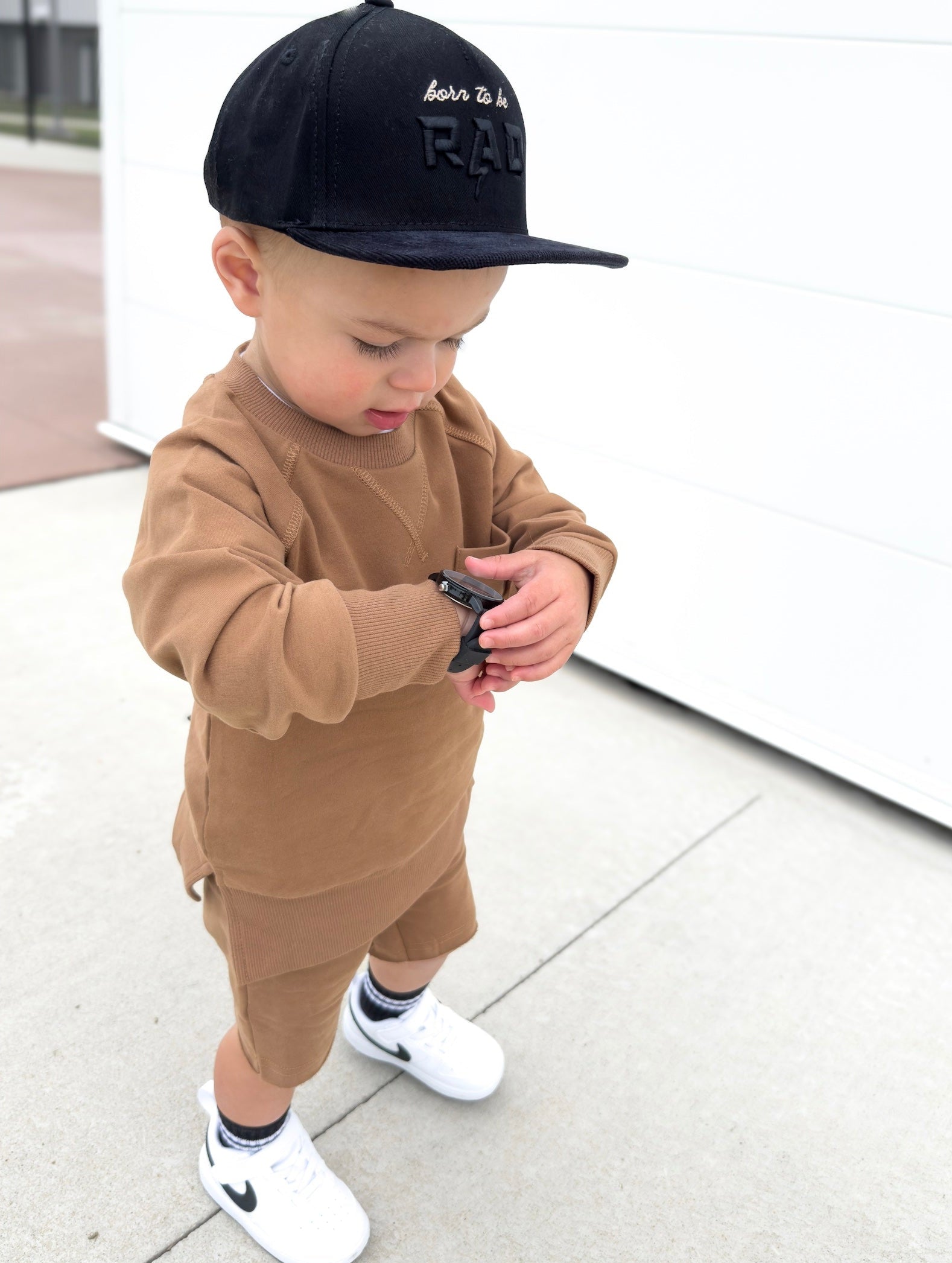 Young boy wearing almond Core Collection toddler set, standing outdoors and showcasing the relaxed, above-the-knee fit checking the time!