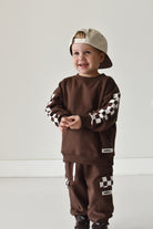 A toddler wearing a matching brown sweatshirt and jogger pants set with black and white checkered sleeve and leg details, along with a beige baseball cap, standing against a plain light gray wall.
