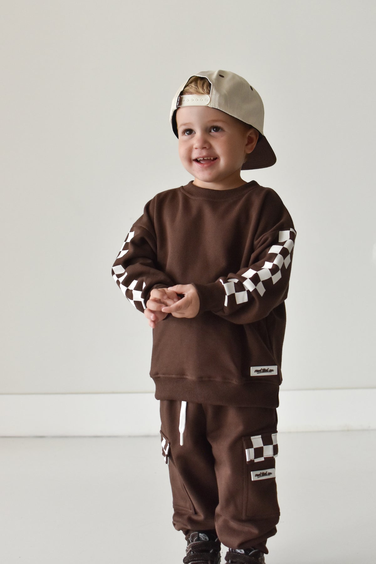 A toddler wearing a matching brown sweatshirt and jogger pants set with black and white checkered sleeve and leg details, along with a beige baseball cap, standing against a plain light gray wall.