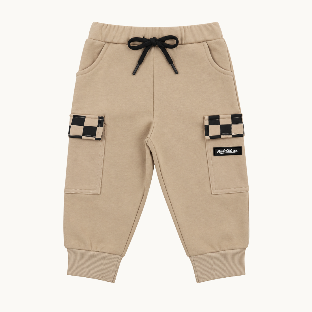 A pair of beige jogger-style cargo pants with a black drawstring, side pockets, and flap cargo pockets featuring a black-and-beige checkerboard pattern. A small black brand patch on the right pocket reads 'rad tod co'.