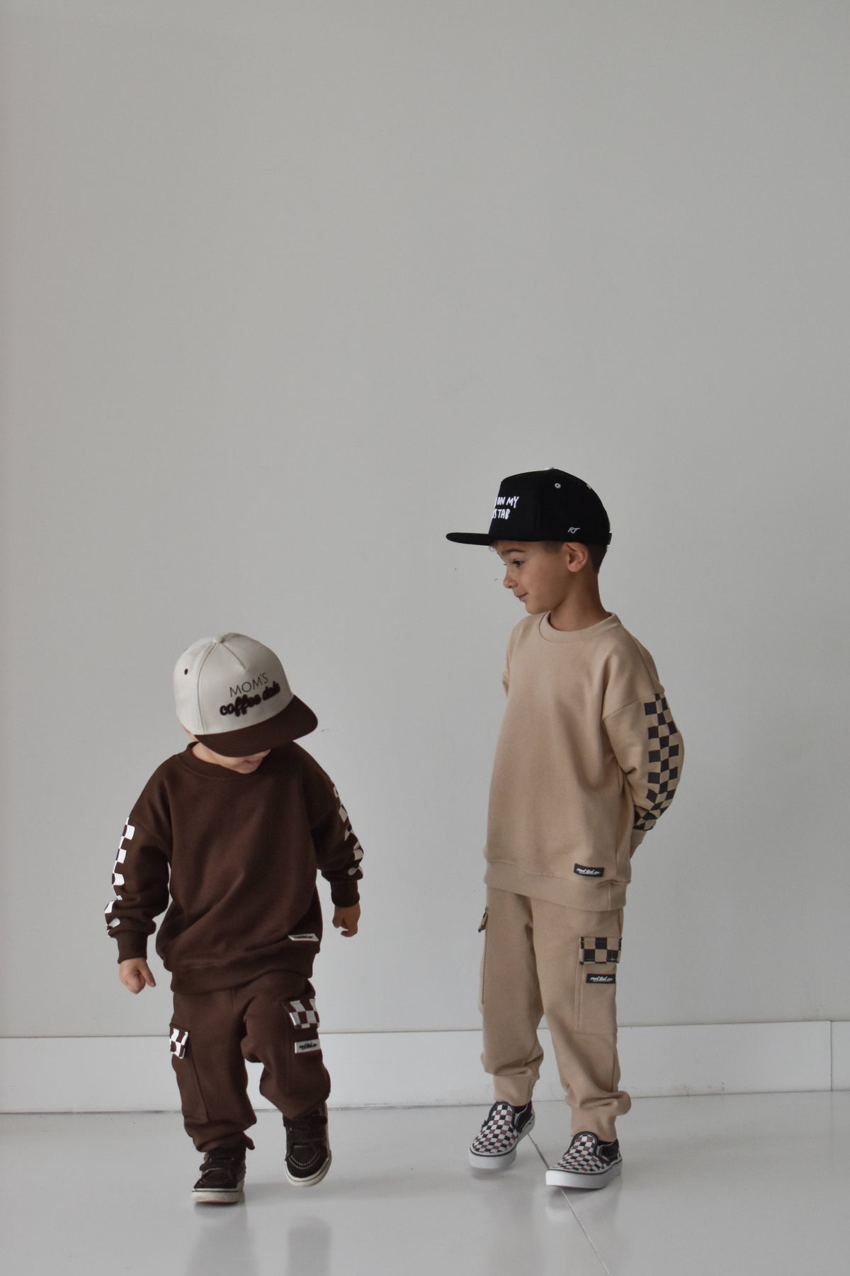 Two children modeling matching streetwear tracksuits in a minimalist studio setting. The child on the left wears a dark brown long-sleeve sweatshirt and matching pants with white graphic details and a white cap.