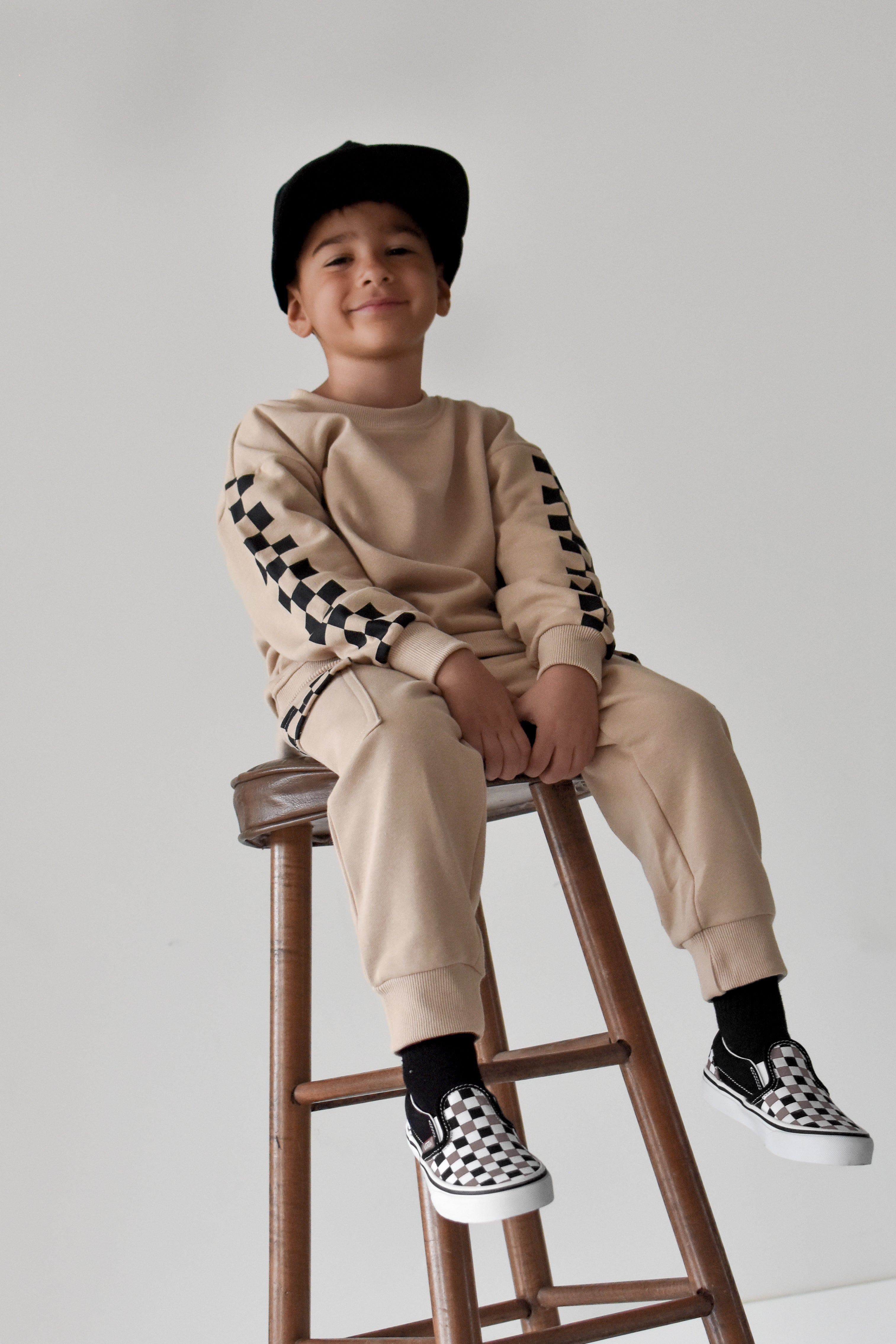 A young boy wearing a beige sweatshirt and jogger pants with black-and-white checkerboard sleeve and sock details, sitting on a wooden stool against a light gray background.