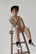 A young boy wearing a beige sweatshirt and jogger pants with black-and-white checkerboard sleeve and sock details, sitting on a wooden stool against a light gray background.