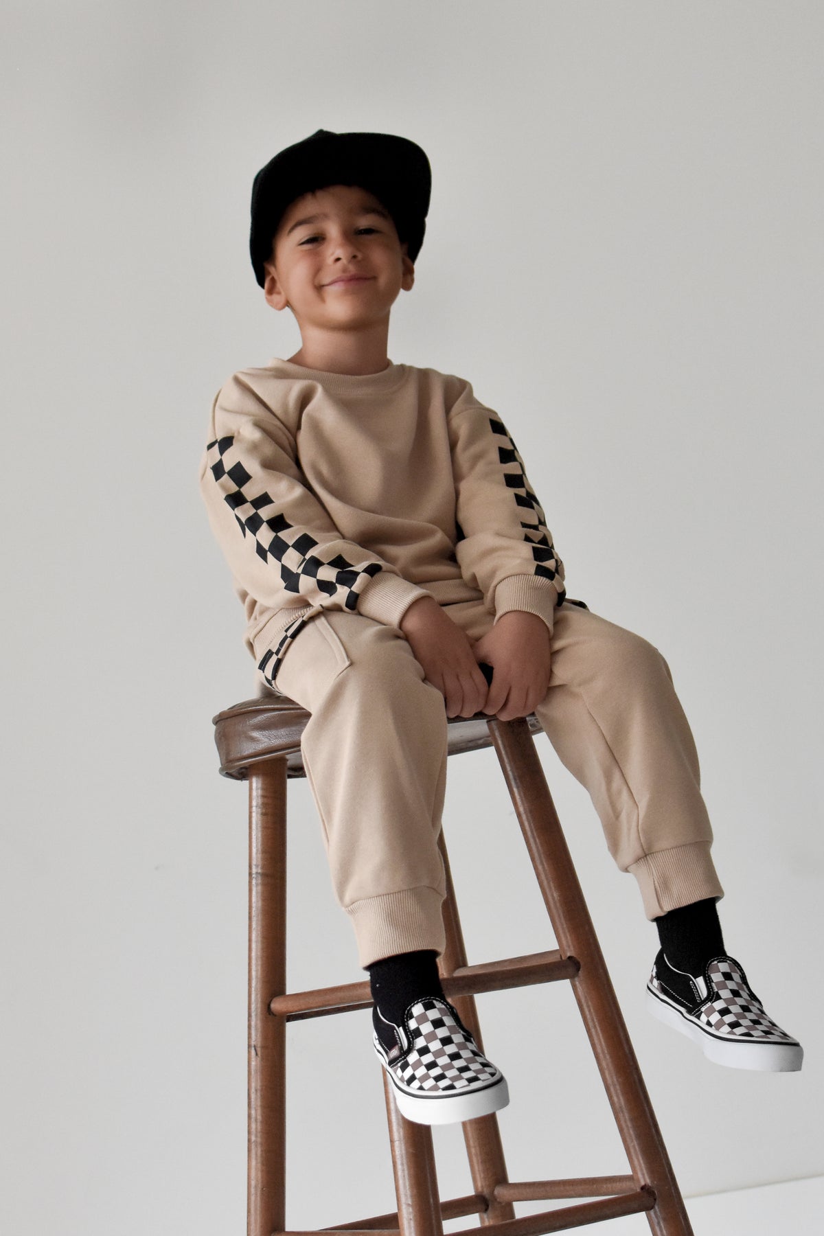 A young boy wearing a beige sweatshirt and jogger pants with black-and-white checkerboard sleeve and sock details, sitting on a wooden stool against a light gray background.