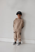 A child stands against a plain white wall wearing a beige sweatshirt and matching sweatpants with black-and-white checkerboard sleeve and leg details, plus a black cap and black-and-white sneakers.