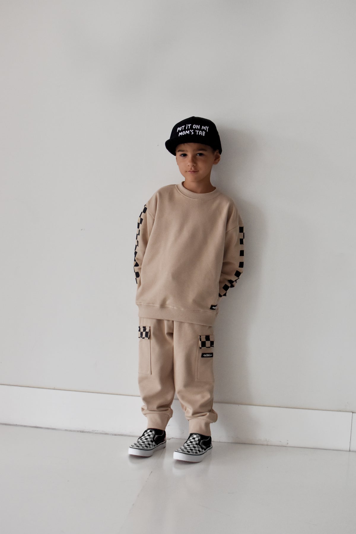A child stands against a plain white wall wearing a beige sweatshirt and matching sweatpants with black-and-white checkerboard sleeve and leg details, plus a black cap and black-and-white sneakers.