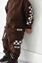 A child wearing a matching brown sweatshirt and jogger pants set with white and black checkerboard accents on the sleeves and cargo pockets, standing against a white wall.