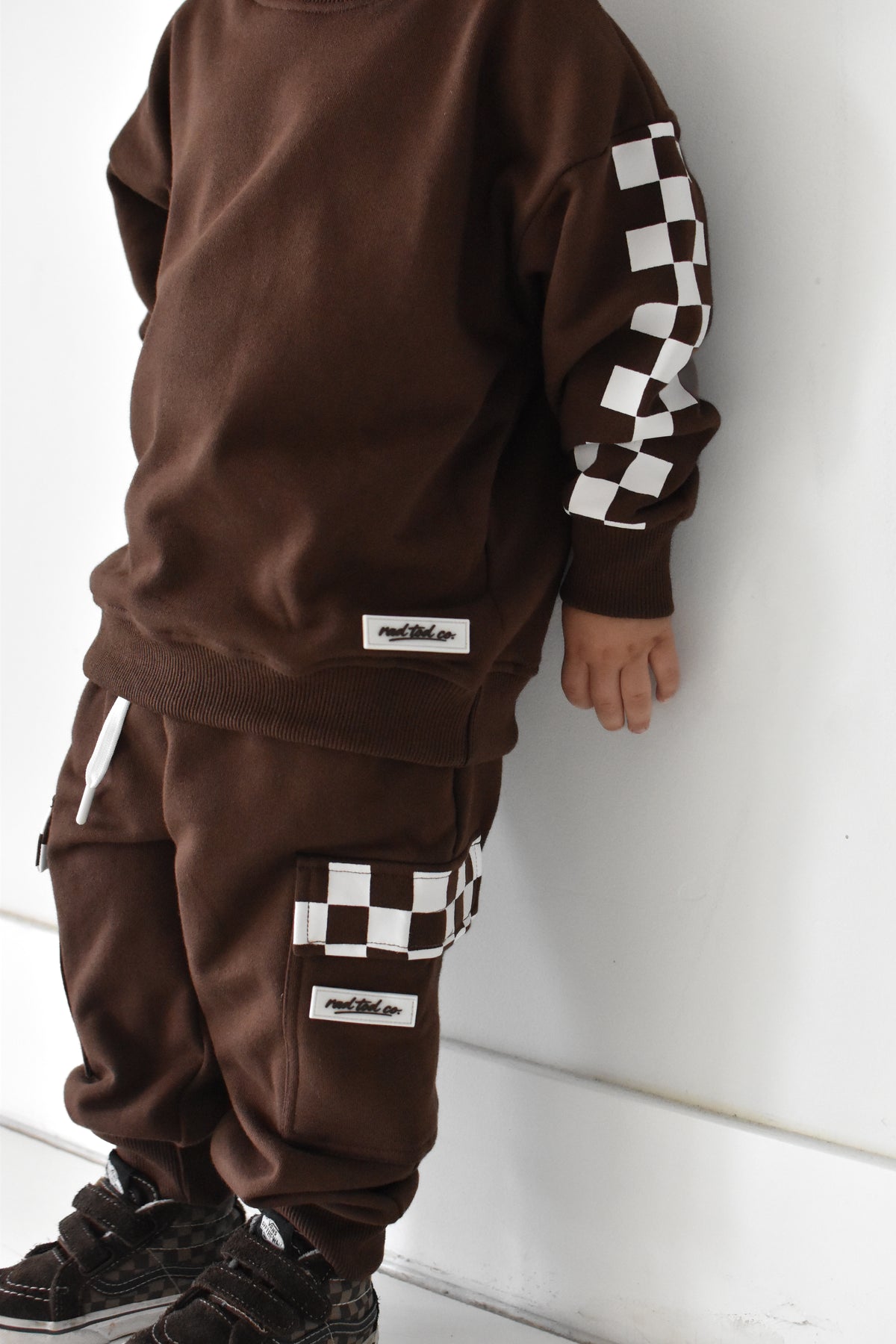 A child wearing a matching brown sweatshirt and jogger pants set with white and black checkerboard accents on the sleeves and cargo pockets, standing against a white wall.