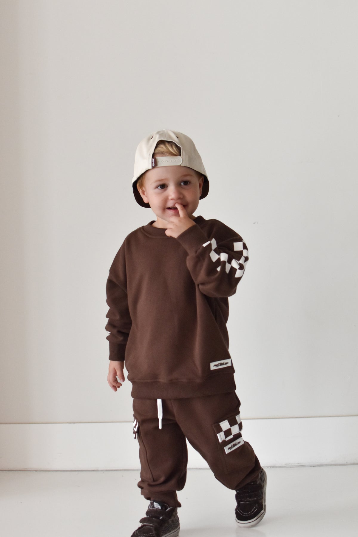 A toddler wearing a matching brown sweatshirt and jogger pants set with white checkerboard accents, a beige baseball cap, and black sneakers, standing against a plain white wall.