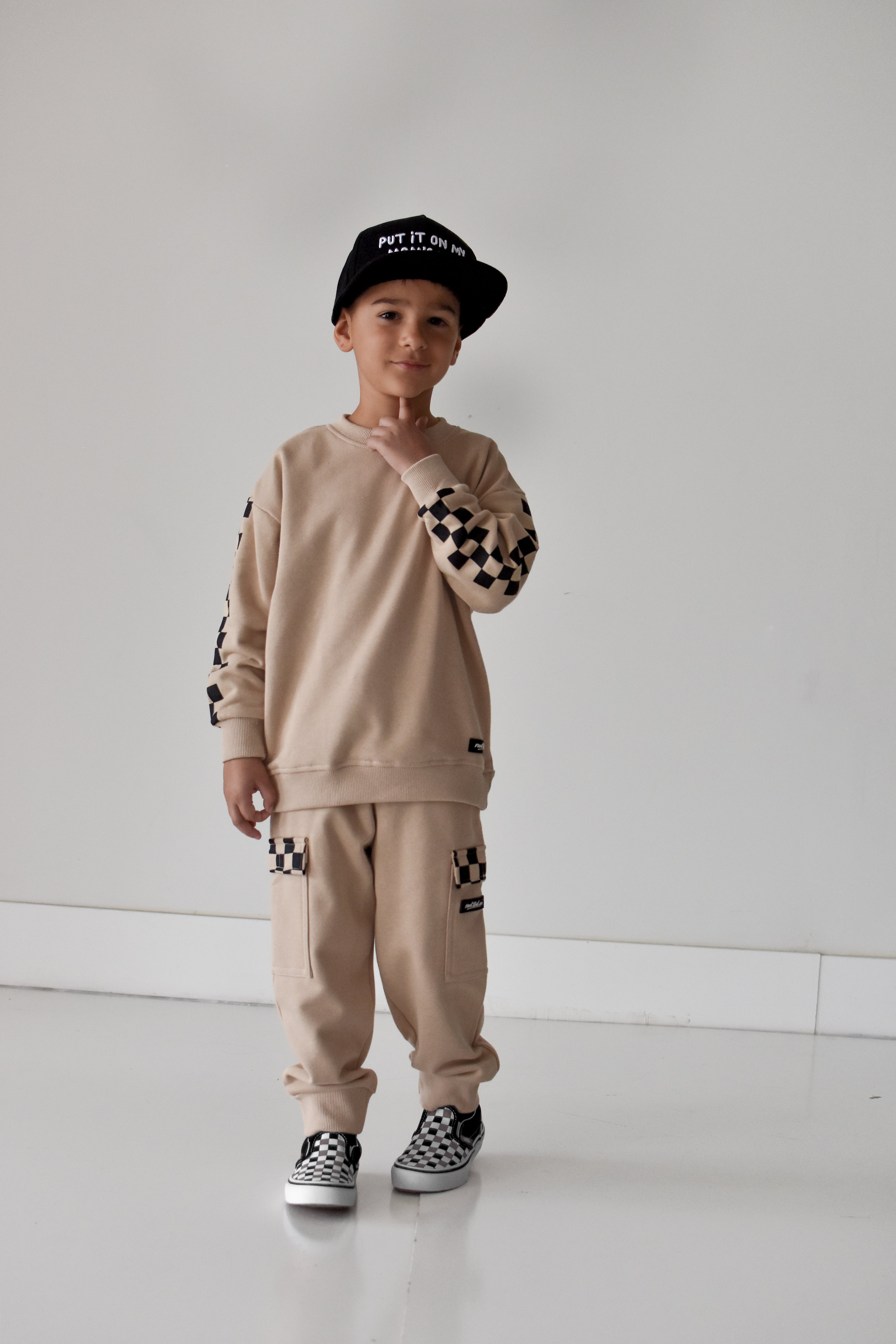 A young boy modeling a beige two-piece tracksuit with black and white checkerboard pattern details on the sleeves, pants, and cap, standing against a plain white studio background.