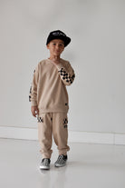 A young boy modeling a beige two-piece tracksuit with black and white checkerboard pattern details on the sleeves, pants, and cap, standing against a plain white studio background.