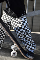 A person wearing black and white checkered cargo pants with a skateboard, captured in an outdoor urban setting.