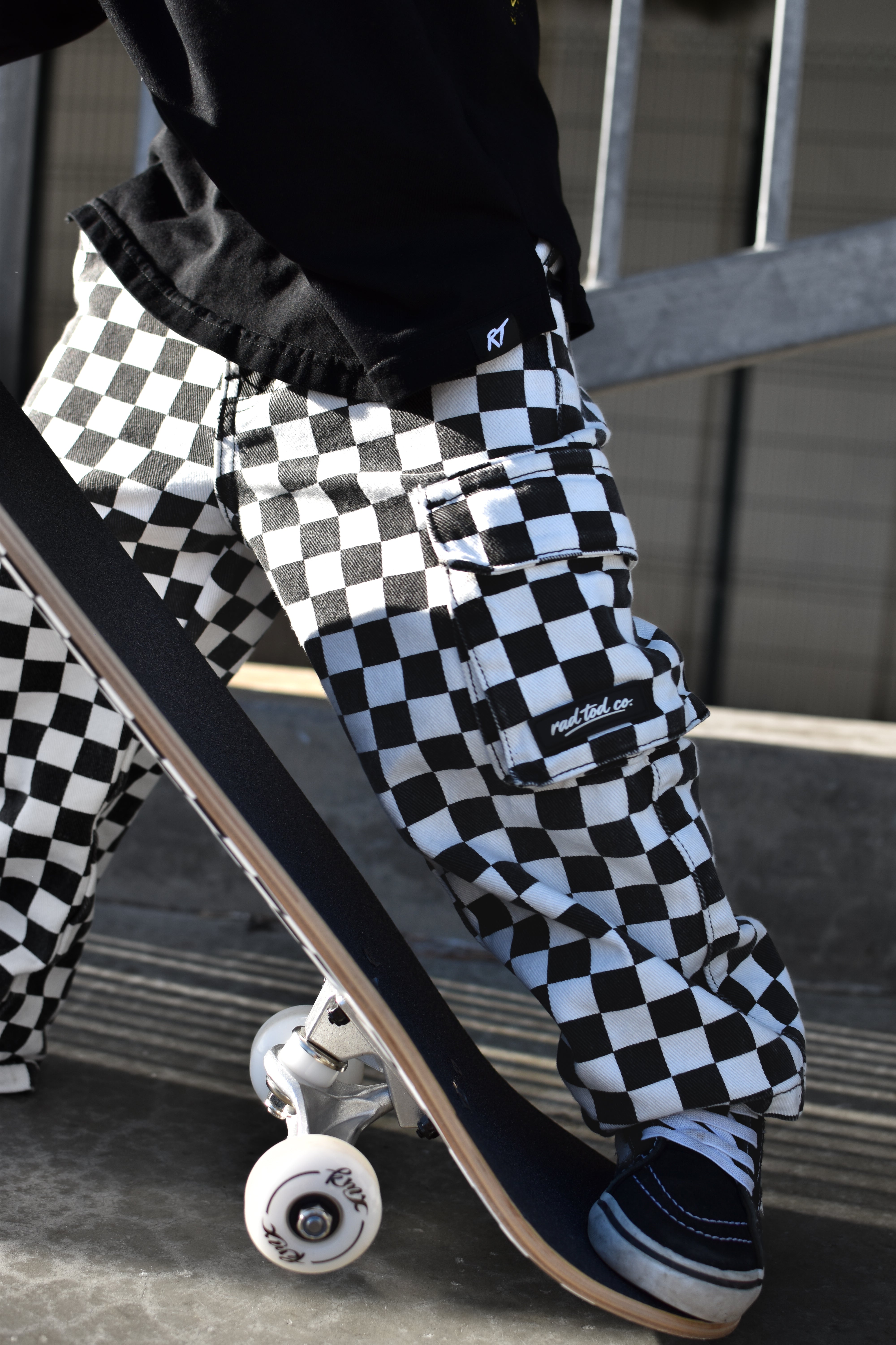 A person wearing black and white checkered cargo pants with a skateboard, captured in an outdoor urban setting.