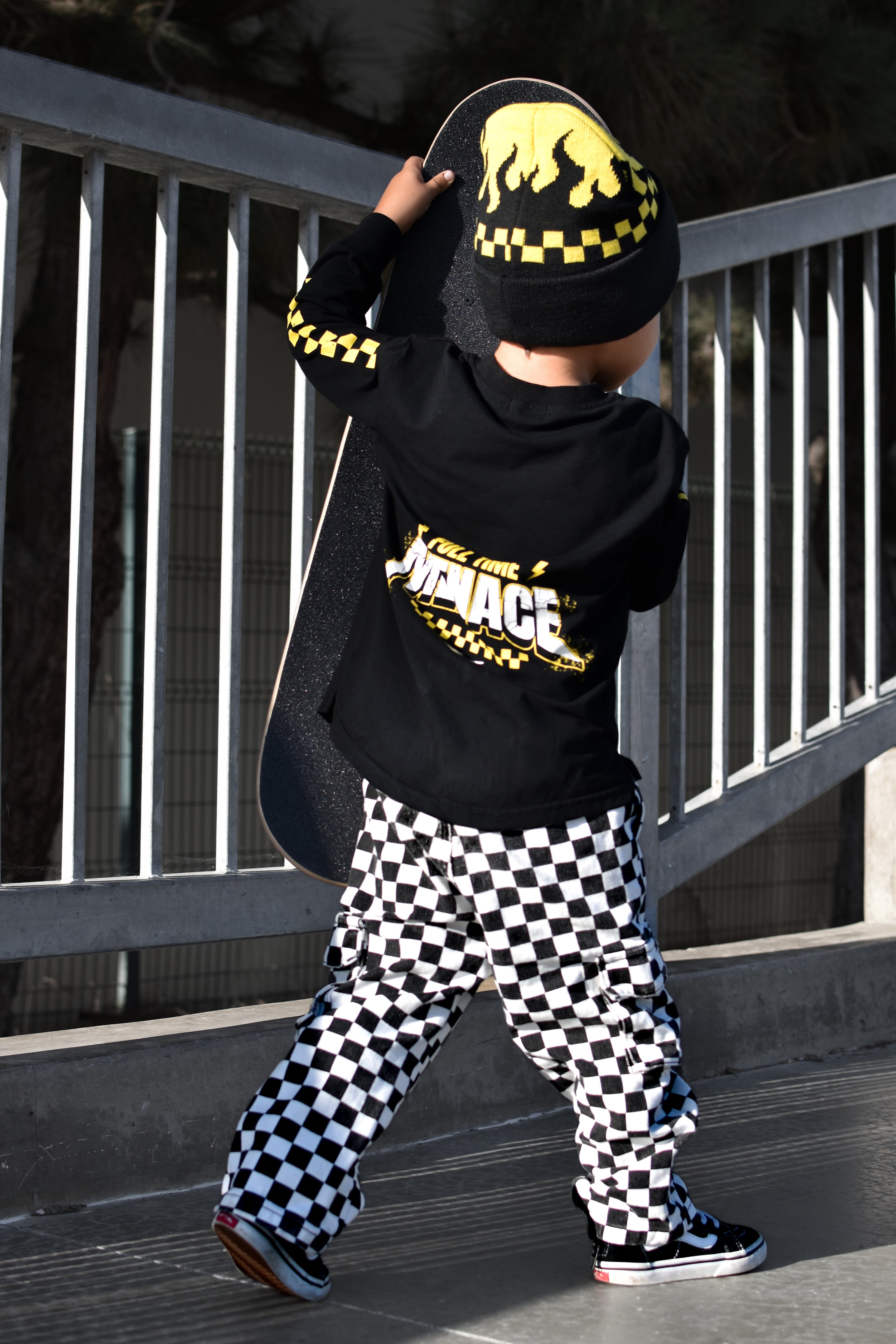 A child wearing a black and yellow checkered beanie, a black long-sleeve top with a yellow and white 'MENACE' graphic on the back, and black-and-white checkered pants, holding a skateboard outdoors near a metal railing.