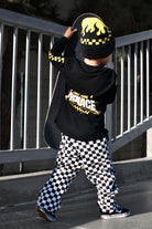 A child wearing a black and yellow checkered beanie, a black long-sleeve top with a yellow and white 'MENACE' graphic on the back, and black-and-white checkered pants, holding a skateboard outdoors near a metal railing.