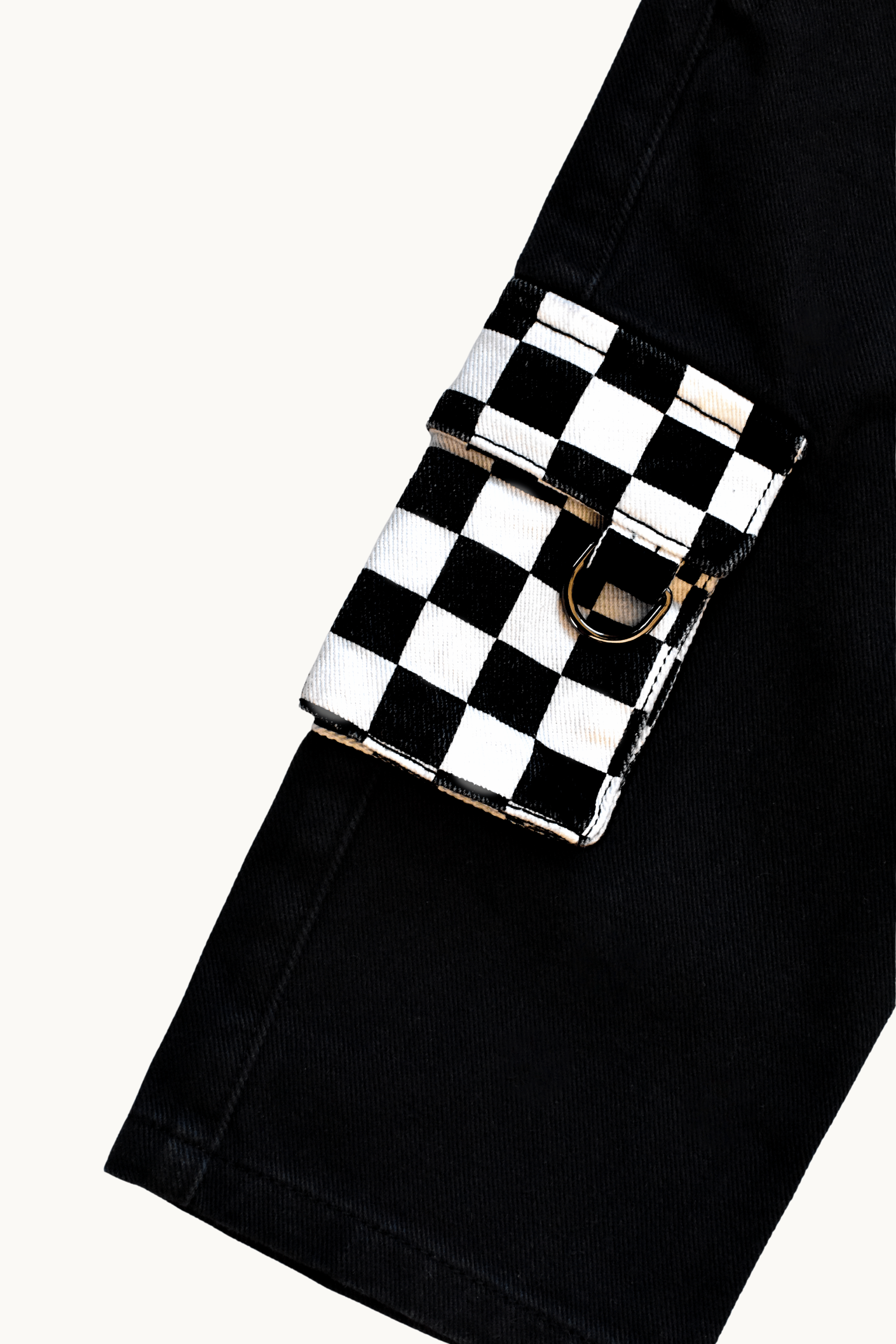 A close-up of a black and white checkerboard flap pouch with a silver-tone metal D-ring, attached to a black denim belt or pants leg, set against a plain off-white background.
