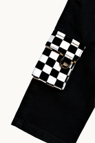 A close-up of a black and white checkerboard flap pouch with a silver-tone metal D-ring, attached to a black denim belt or pants leg, set against a plain off-white background.
