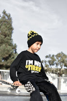 A young boy wearing a black graphic sweatshirt, black beanie with yellow accents, and black checkered pants, sitting on a skateboard at an outdoor skatepark.