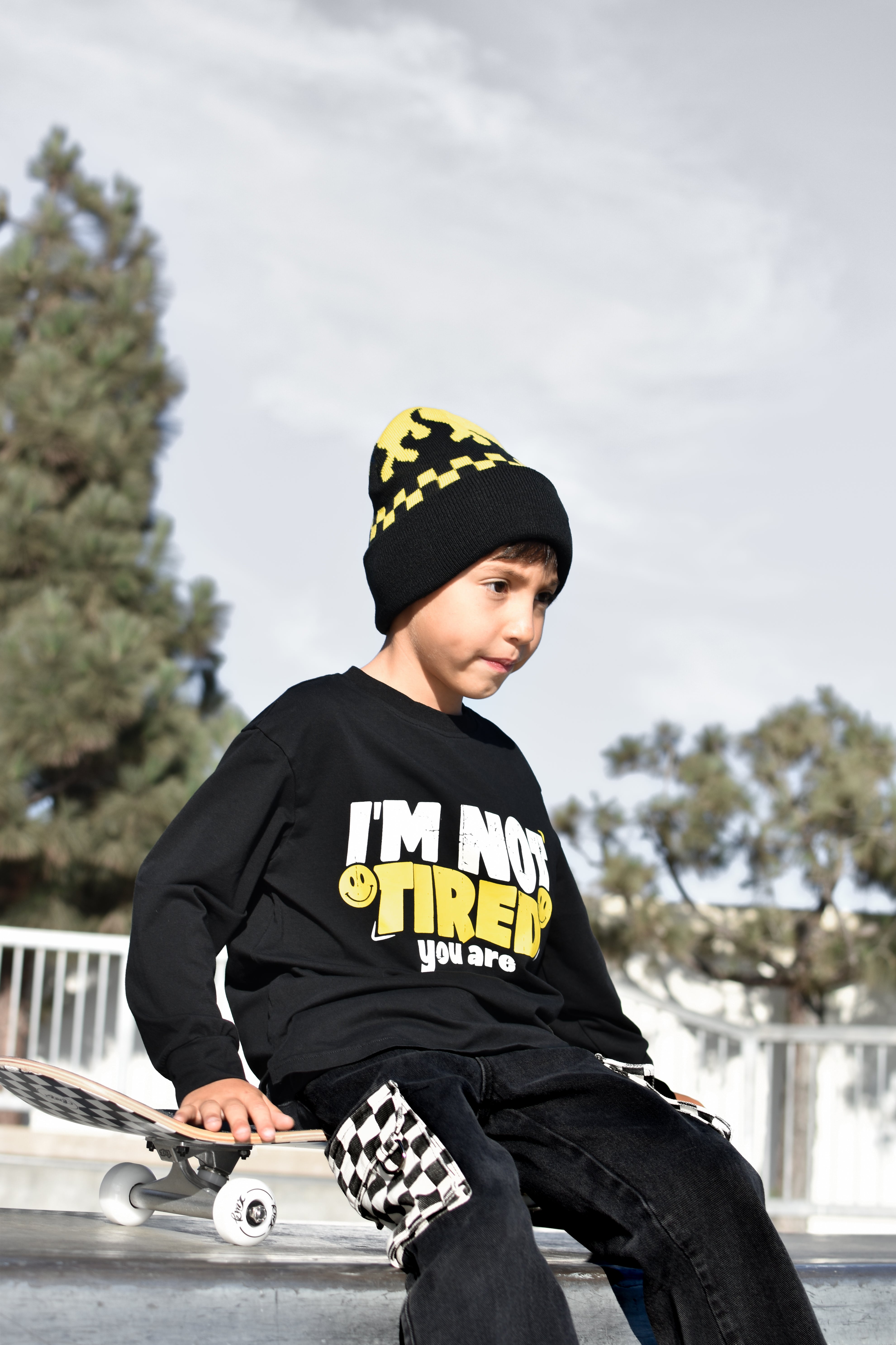 A young boy wearing a black graphic sweatshirt, black beanie with yellow accents, and black checkered pants, sitting on a skateboard at an outdoor skatepark.