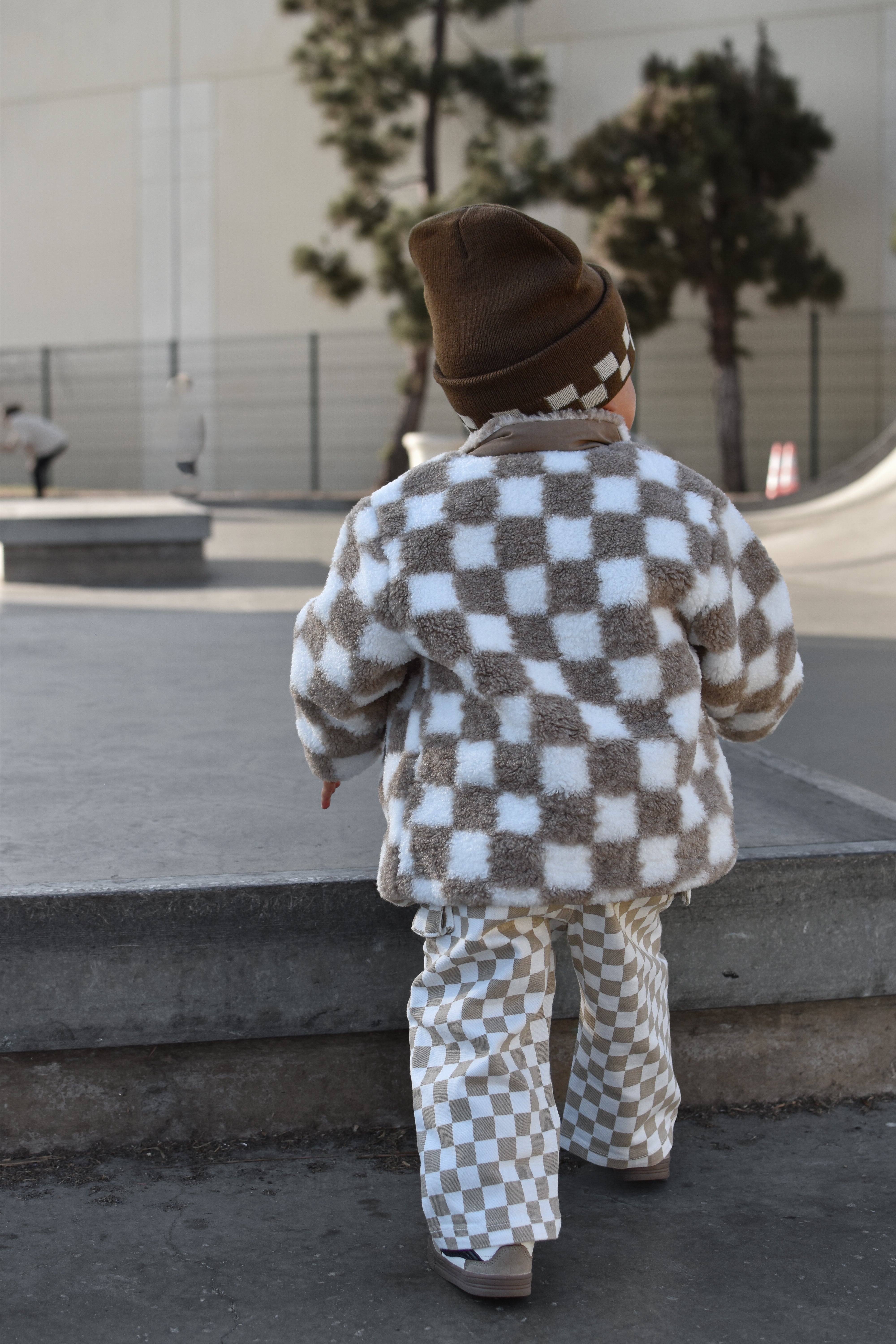 A toddler seen from behind wearing a brown and white checkerboard fleece jacket and matching pants, walking outdoors in a skatepark setting.