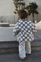 A toddler seen from behind wearing a brown and white checkerboard fleece jacket and matching pants, walking outdoors in a skatepark setting.