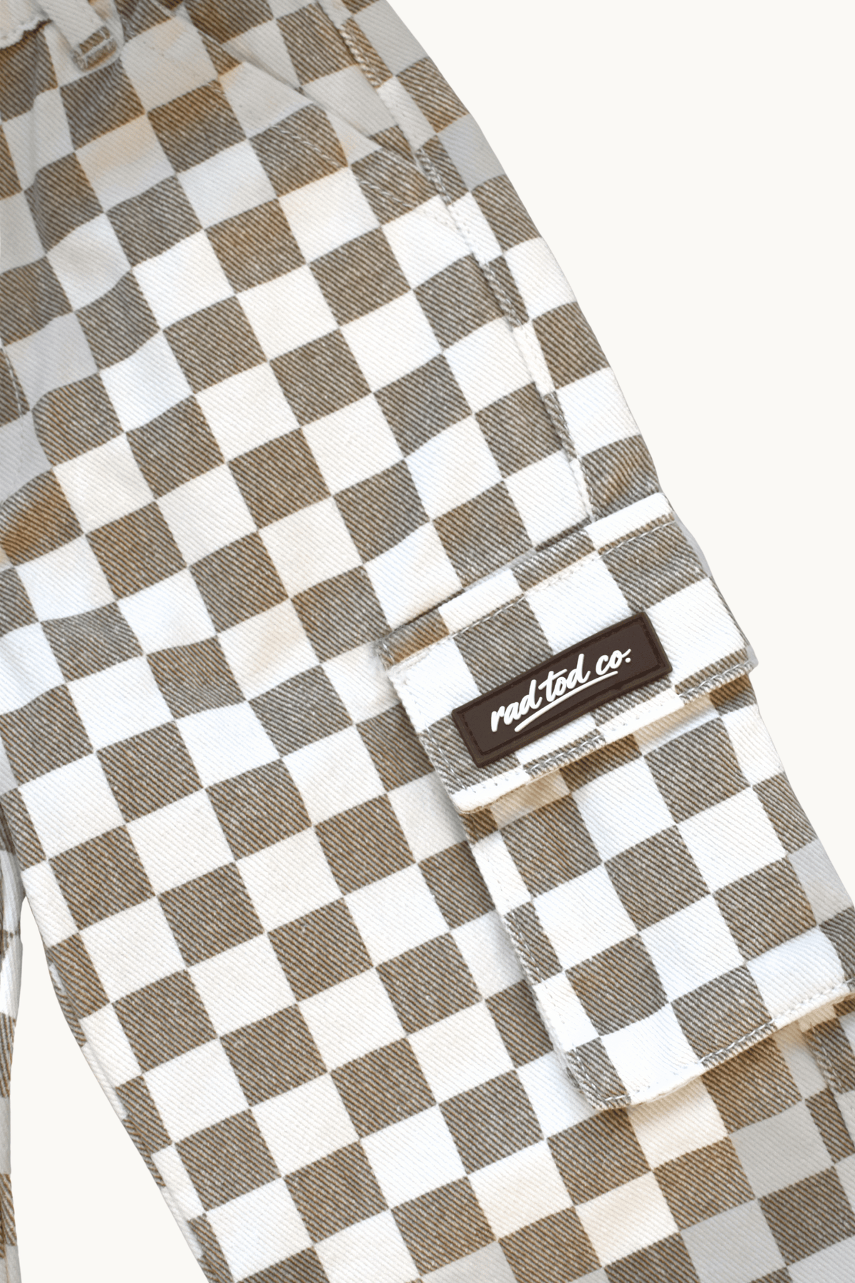 Close-up of a checkered cargo pocket in a brown and white checkerboard pattern, featuring a black brand patch that reads 'Rad Tod co.' on a light off-white background.
