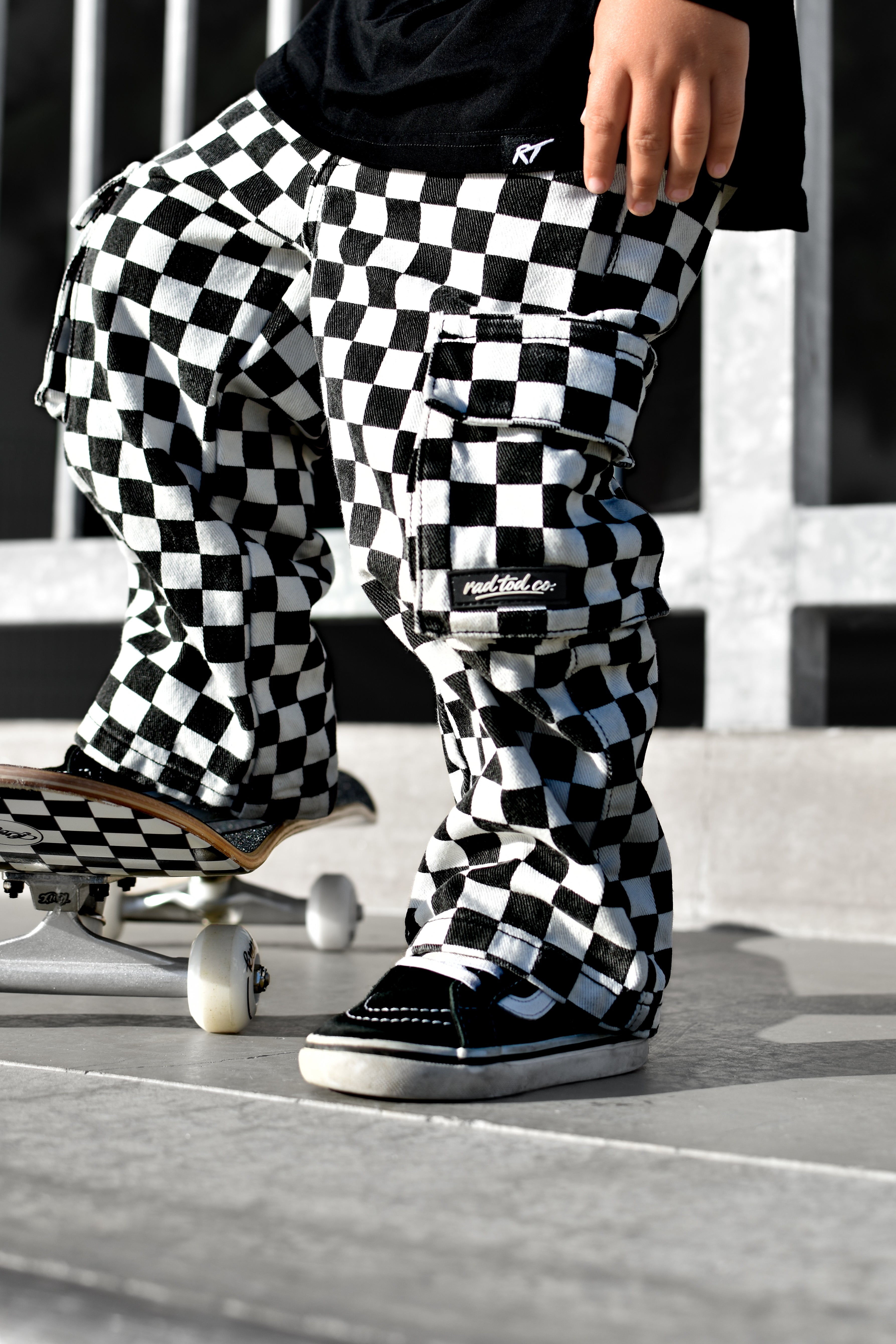 A person wearing black and white checkered cargo pants and black slip-on shoes stands on concrete next to a skateboard. The pants have a bold black-and-white checkerboard pattern and a visible 'Rad Tod CO. ' brand patch on the pocket.