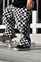 A person wearing black and white checkered cargo pants and black slip-on shoes stands on concrete next to a skateboard. The pants have a bold black-and-white checkerboard pattern and a visible 'Rad Tod CO. ' brand patch on the pocket.