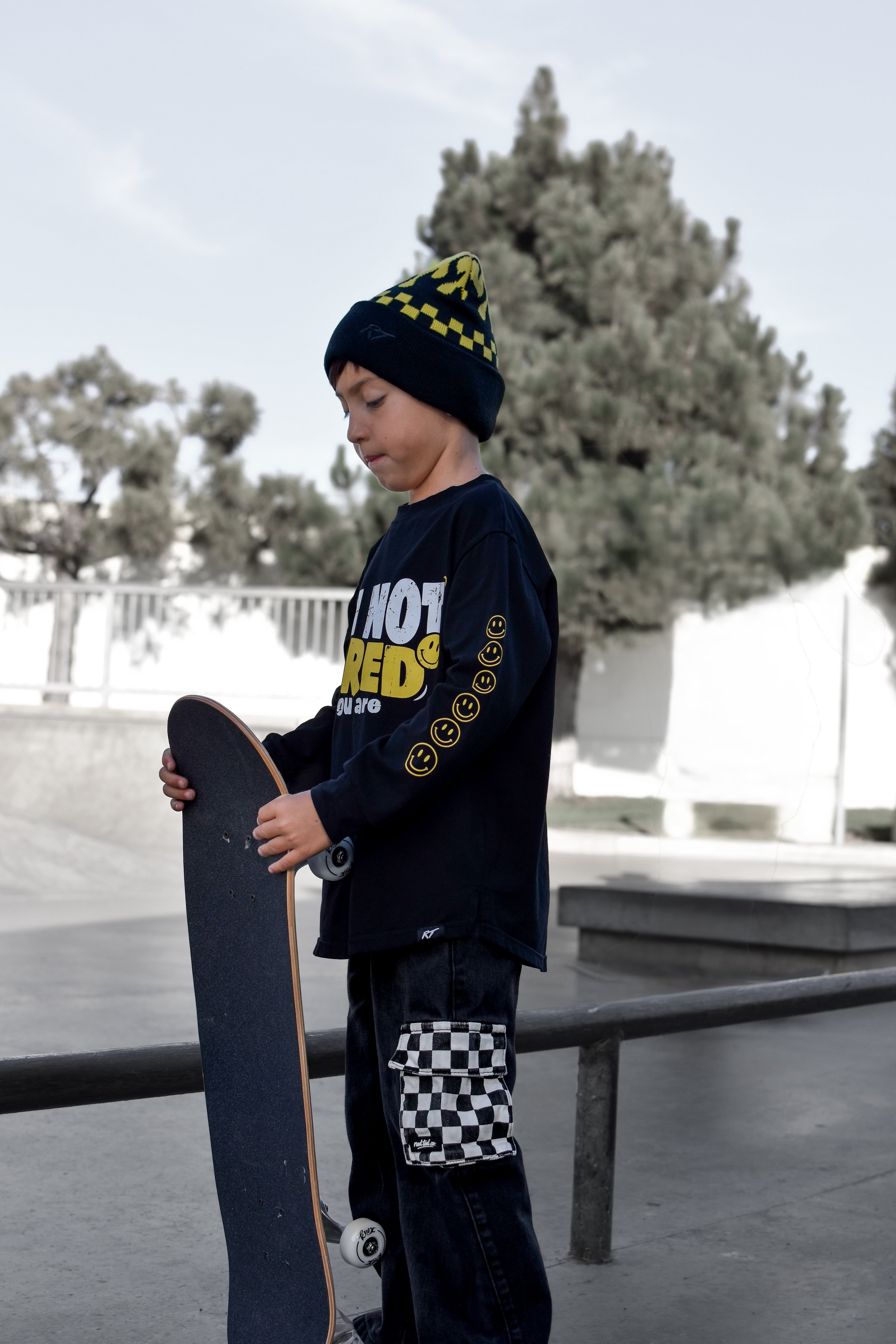 A child standing at a skatepark holding a skateboard, wearing a black beanie with yellow checkered trim, a black long-sleeve graphic shirt with yellow text, and black pants with a checkered patch on the lower leg.