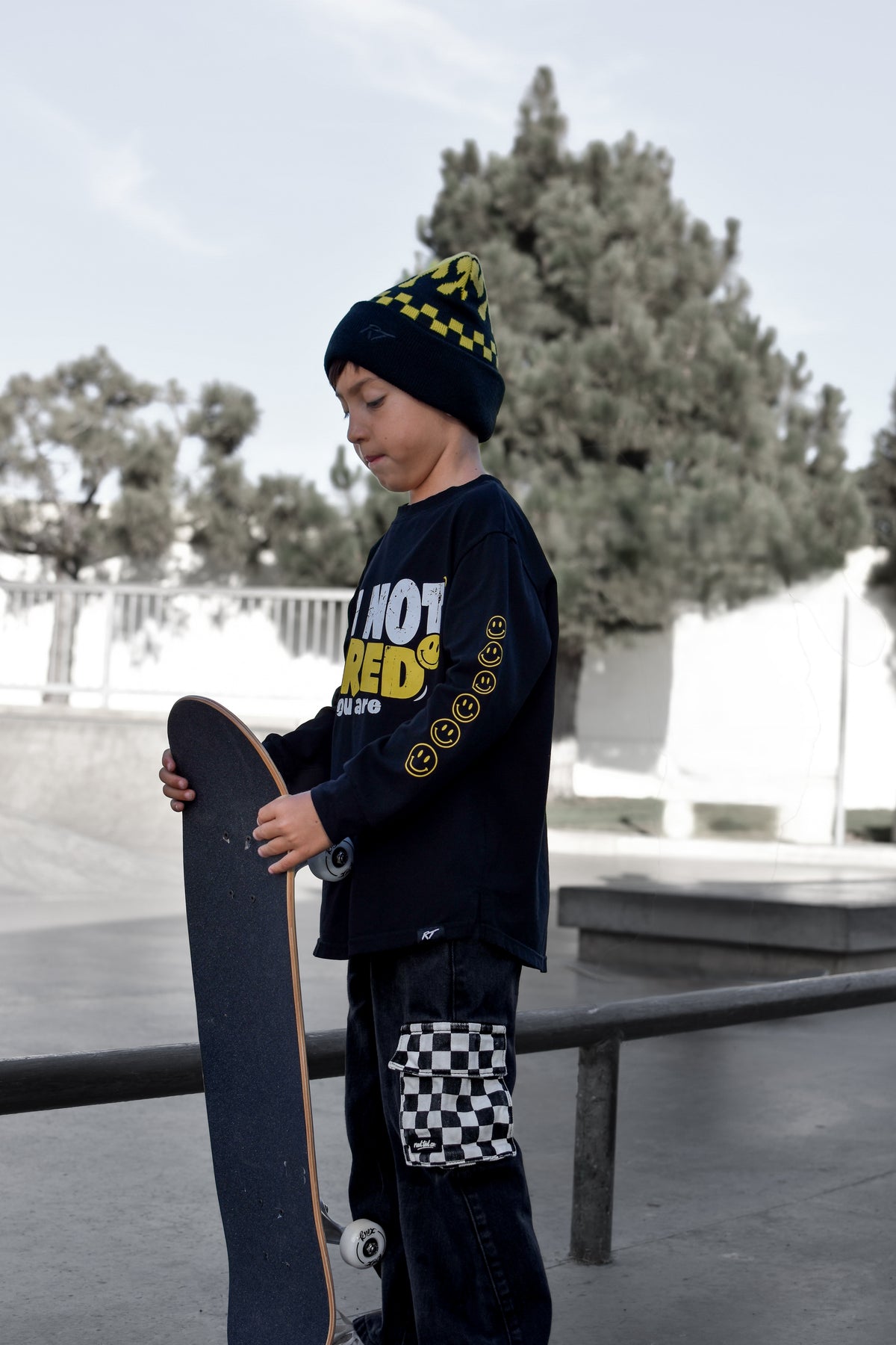 A child standing at a skatepark holding a skateboard, wearing a black beanie with yellow checkered trim, a black long-sleeve graphic shirt with yellow text, and black pants with a checkered patch on the lower leg.