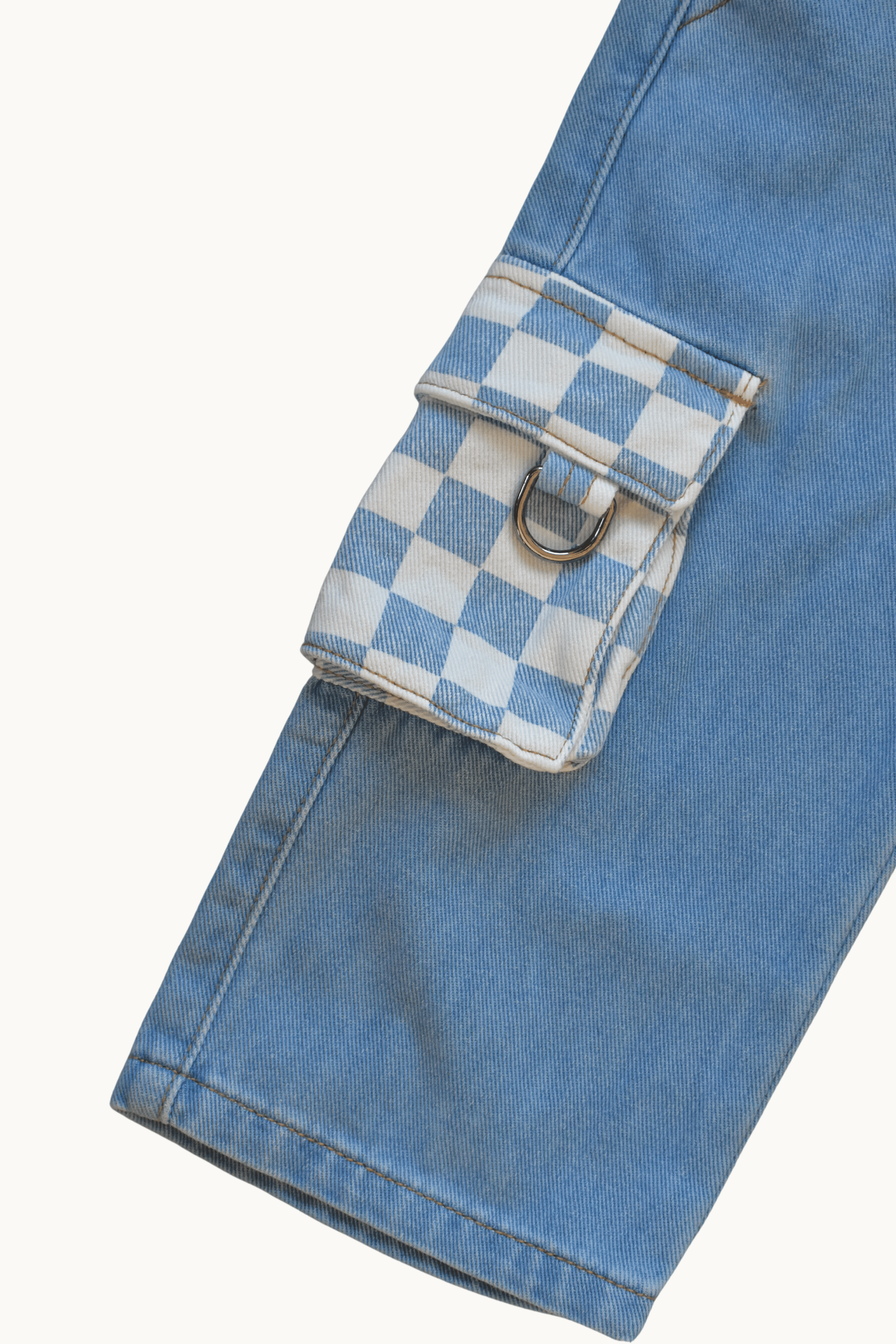 Close-up of a light blue denim jacket or pants leg showing a flap cargo pocket with a white and blue checkered pattern and a silver metal D-ring, on a plain off-white background.