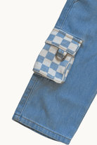 Close-up of a light blue denim jacket or pants leg showing a flap cargo pocket with a white and blue checkered pattern and a silver metal D-ring, on a plain off-white background.